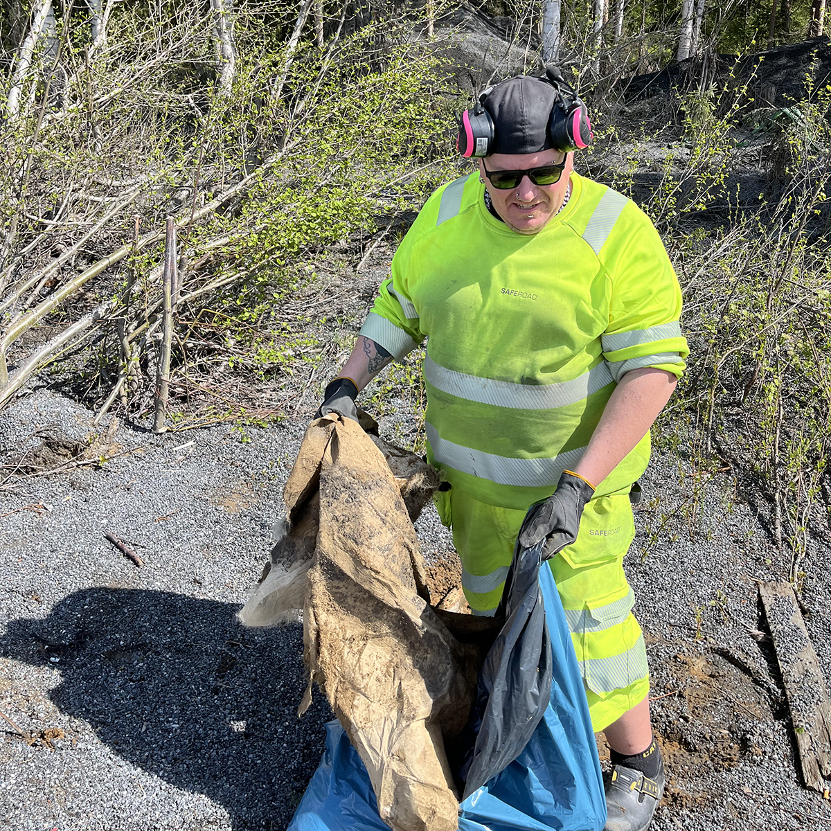Man i varselkläder plockar skräp i naturen och lägger i en blå sop-påse