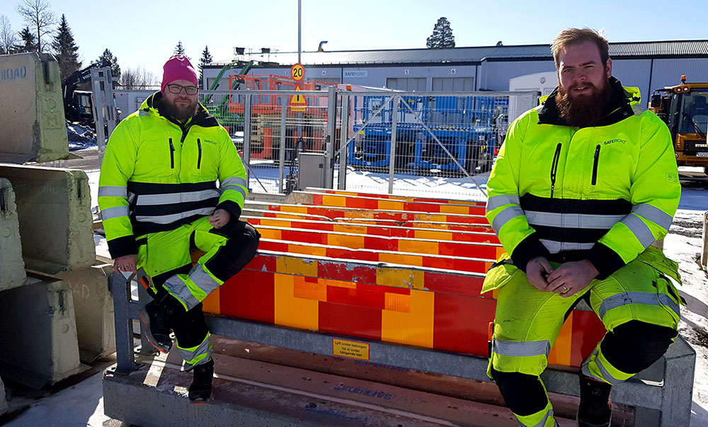 Två killar poserar framför TA-balk i varselkläder