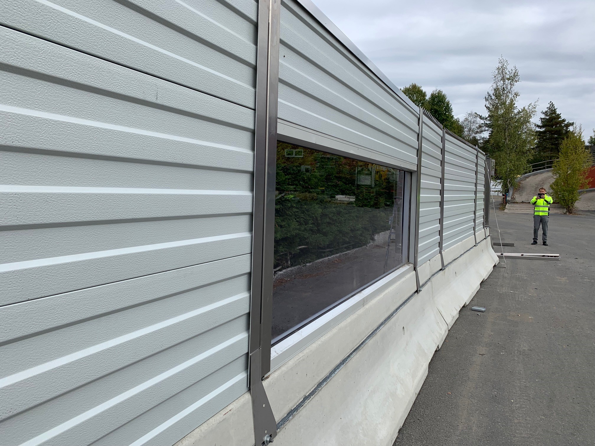 Grå bullerskärm med ett fönster i mitten, som är monterad på skyddsbarriär 